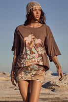 Woman wearing a brown graphic t-shirt and patterned skirt on a beach.