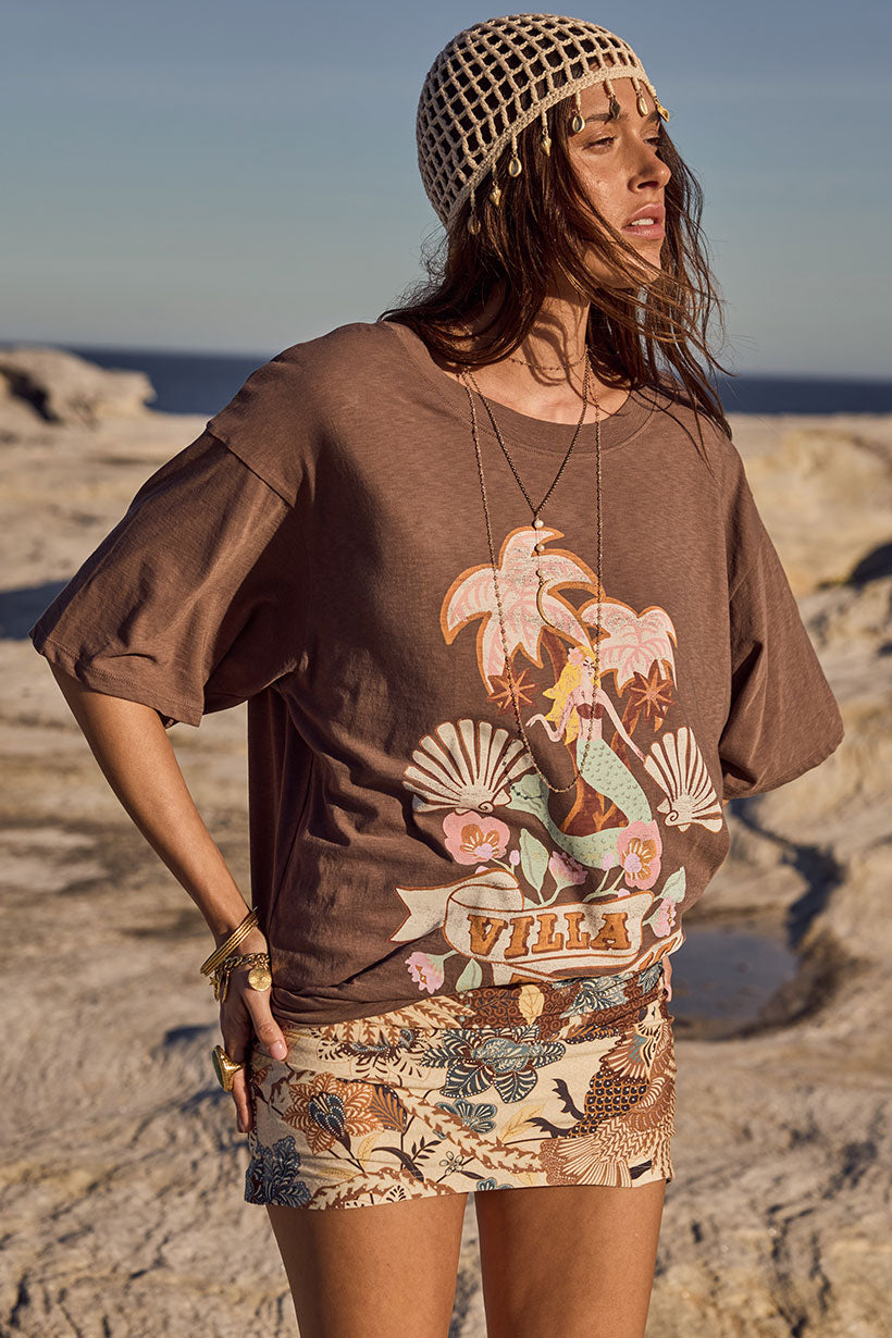 Woman wearing a brown graphic t-shirt with a mermaid design on a rocky beach.