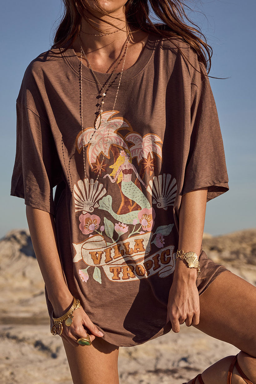Person wearing a brown t-shirt with a mermaid graphic design in a desert setting