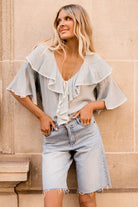 Woman wearing a light blue blouse with ruffled sleeves and denim shorts against a beige wall.