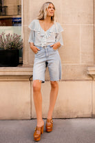 Woman wearing a light blue blouse with ruffles, denim shorts, and brown clogs standing against a beige wall.