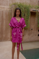 Woman wearing a pink dress with palm tree designs against a beige wall with greenery.
