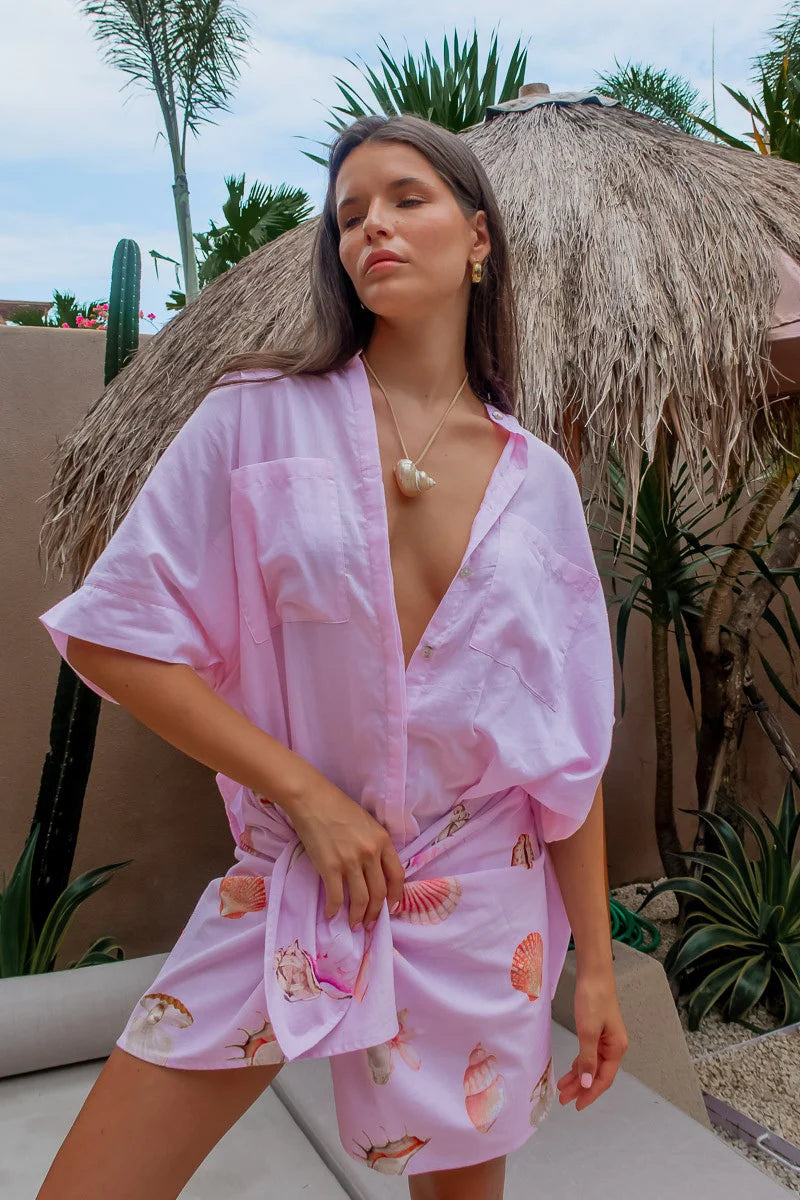 Woman wearing a pink robe with shell patterns under a thatched umbrella.