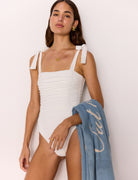Woman wearing a white swimsuit with a blue towel draped over her shoulder against a light background