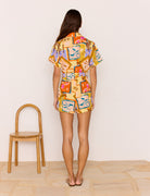 Woman wearing a colorful patterned outfit standing in a minimal room with a wooden chair.