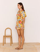 Woman wearing a colorful patterned outfit standing next to a wooden chair against a plain background