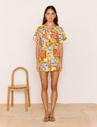 Woman wearing a colorful patterned outfit standing next to a wooden chair on a light wood floor.