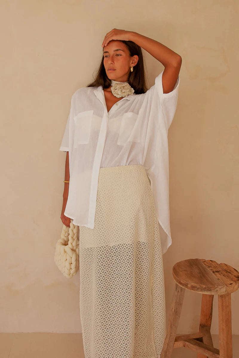Woman wearing a white shirt and beige skirt against a beige wall with a wooden stool.