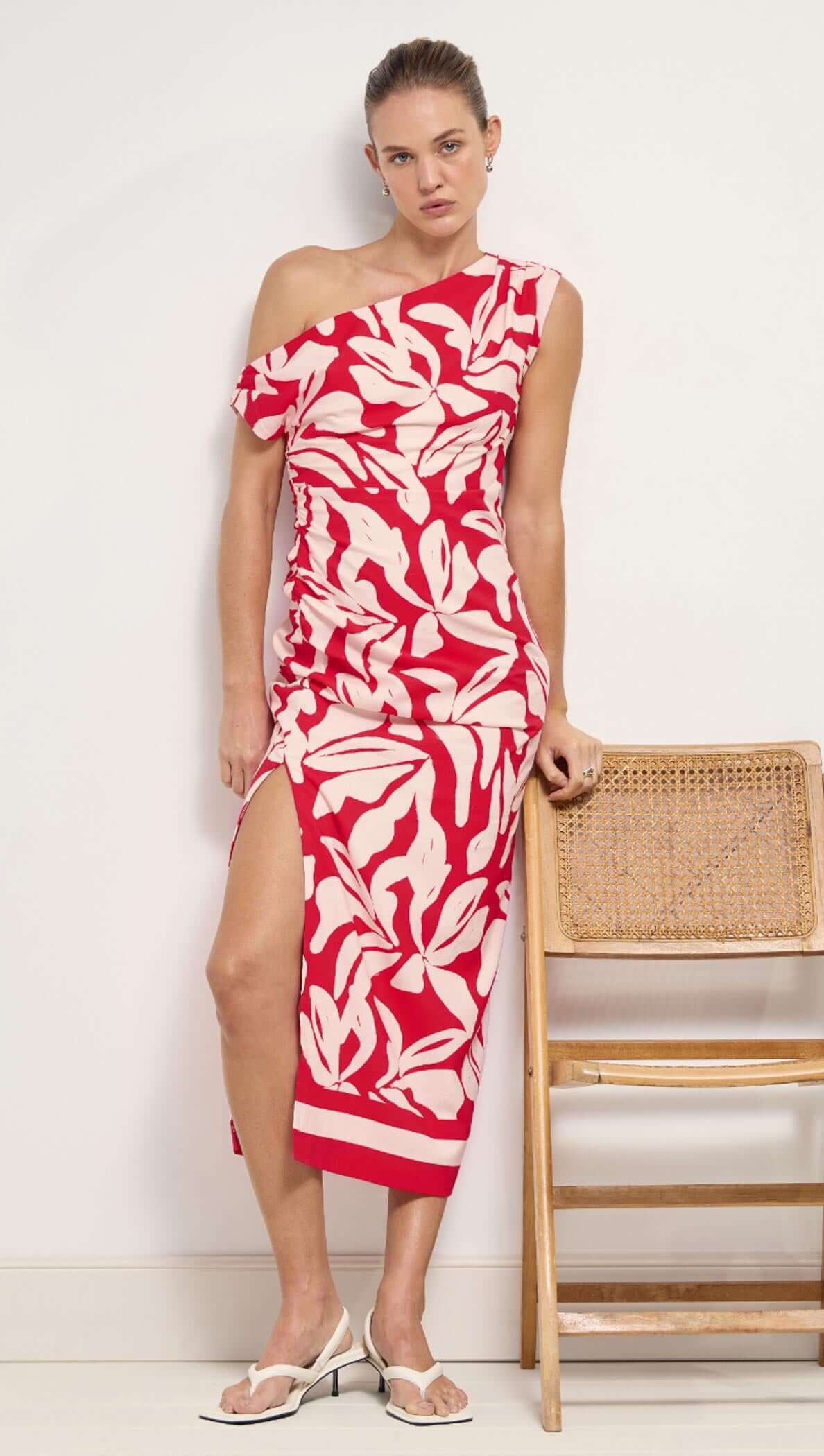 Woman wearing a red and white patterned dress standing next to a wooden chair against a white wall.