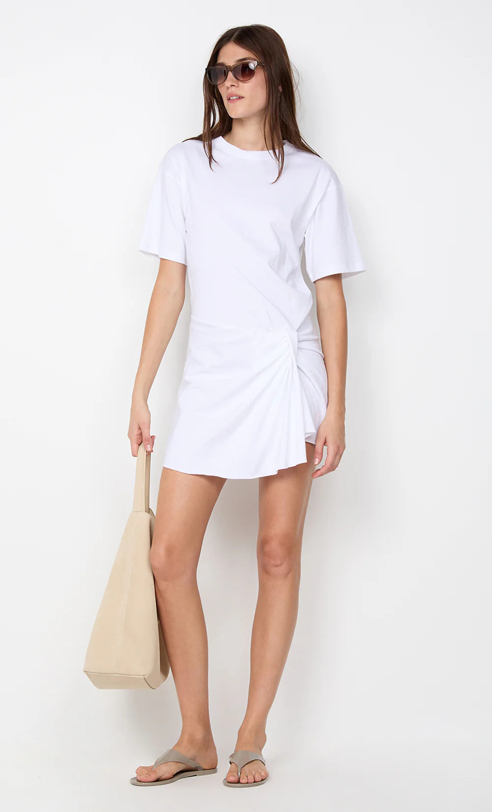 Woman wearing a white t-shirt dress holding a beige tote bag on a white background
