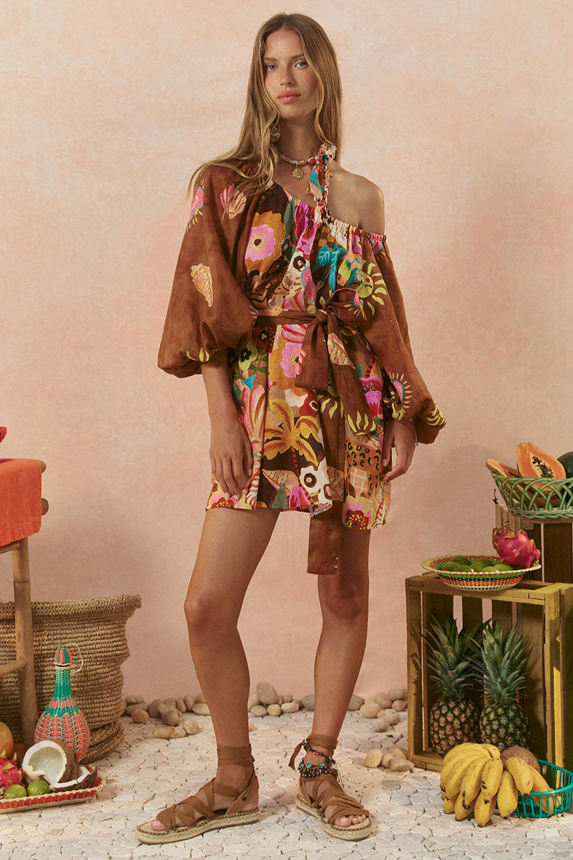A model wearing a sleeveless tunic dress with a floral pattern in a tan color, standing in a setting with tropical fruits and decorative items.