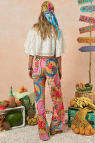 Person wearing colorful pants and a headscarf in a room with decorative items and fruit.