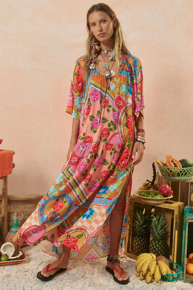 Woman wearing a colorful floral dress standing in a decorative setting with fruits and a pink wall.