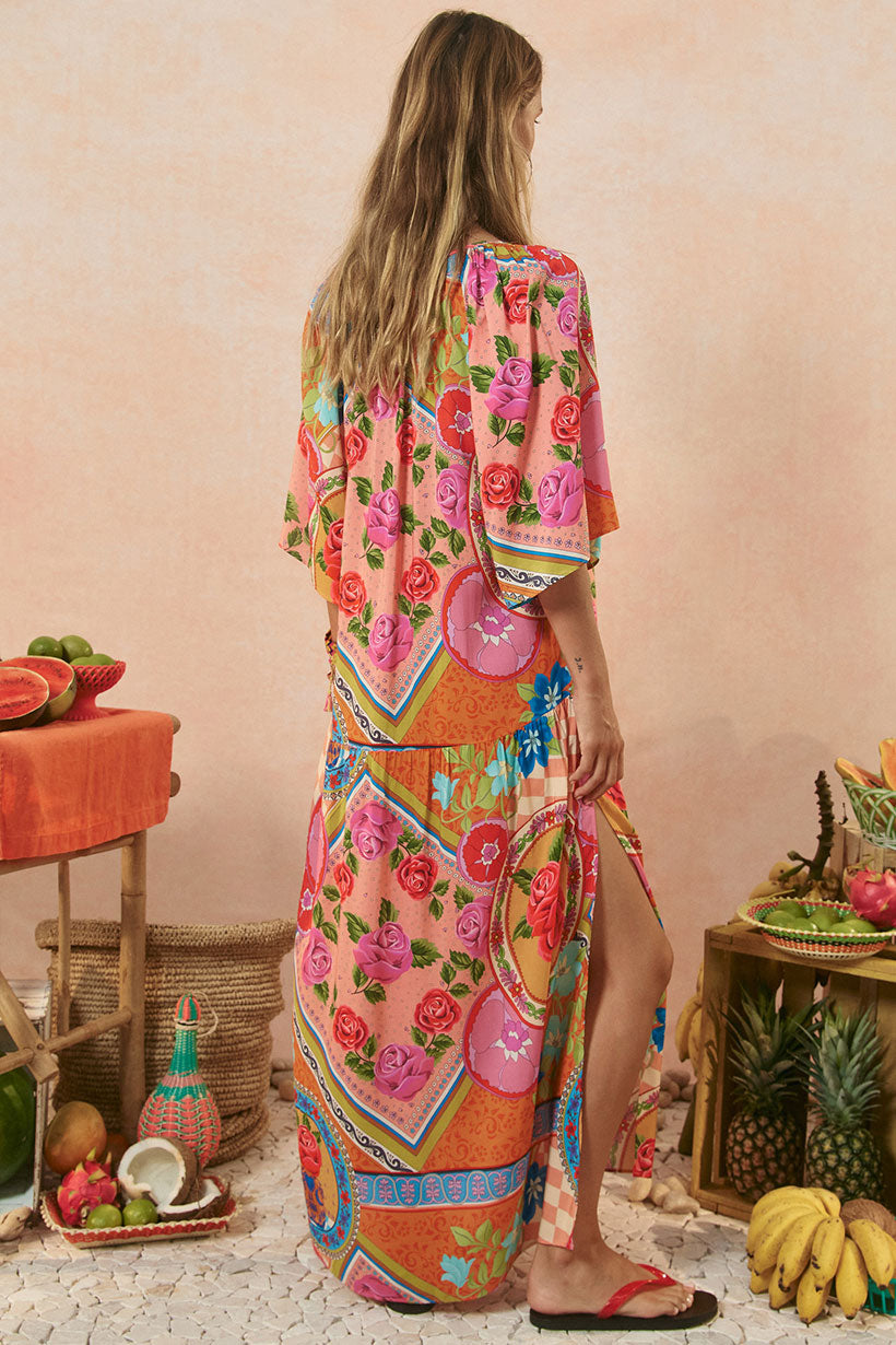 Woman wearing a colorful floral dress standing in a tropical setting with fruits and plants.