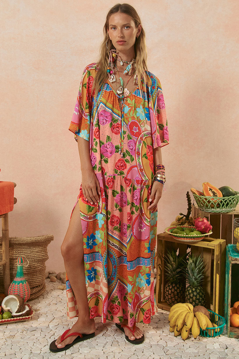 Woman wearing a colorful patterned dress standing in a decorative setting with fruits and plants.
