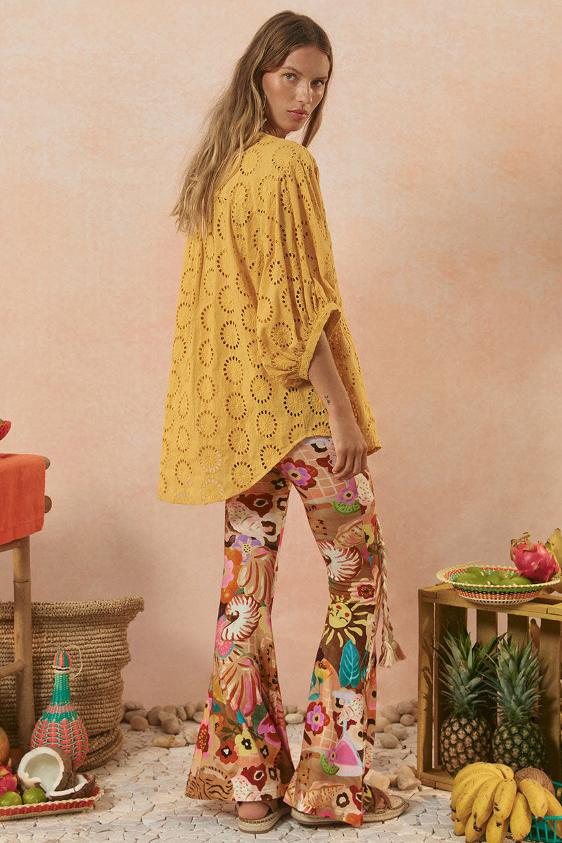 Woman wearing a yellow crochet top and floral pants standing in a decorative setting with fruits and plants.