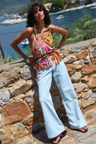 Woman in a colorful top and light blue jeans standing against a stone wall with a scenic background.