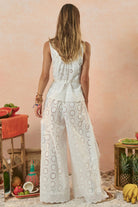 Woman wearing a white lace jumpsuit standing in a tropical setting with fruits and a pink wall.