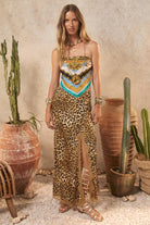 Woman in a leopard print skirt with a scarf around her neck, standing in a desert-like setting with cacti and pots.