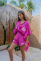 Woman in a pink outfit with floral patterns standing under a thatched umbrella in a tropical setting.