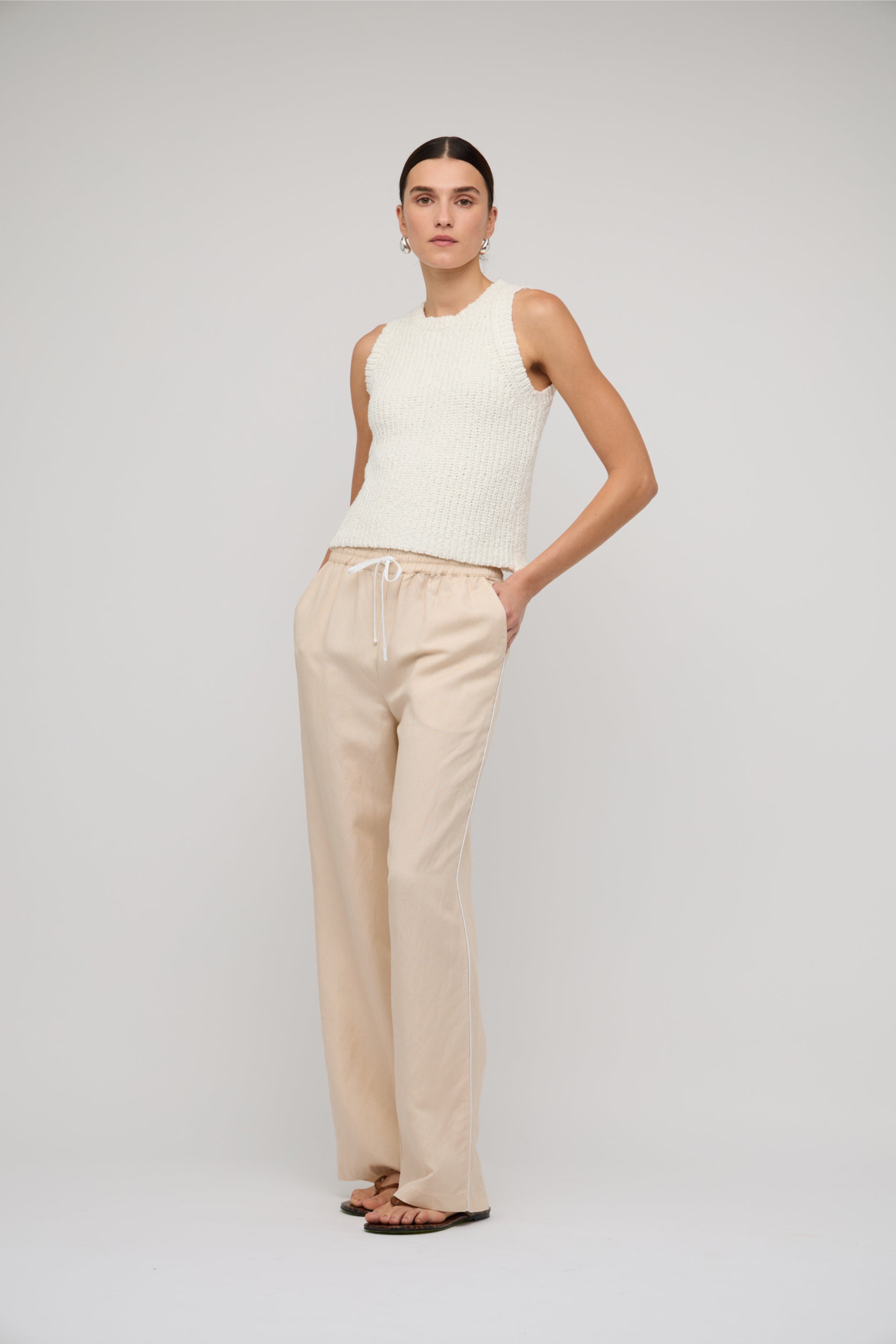 Woman wearing a white sleeveless top and beige pants on a plain background