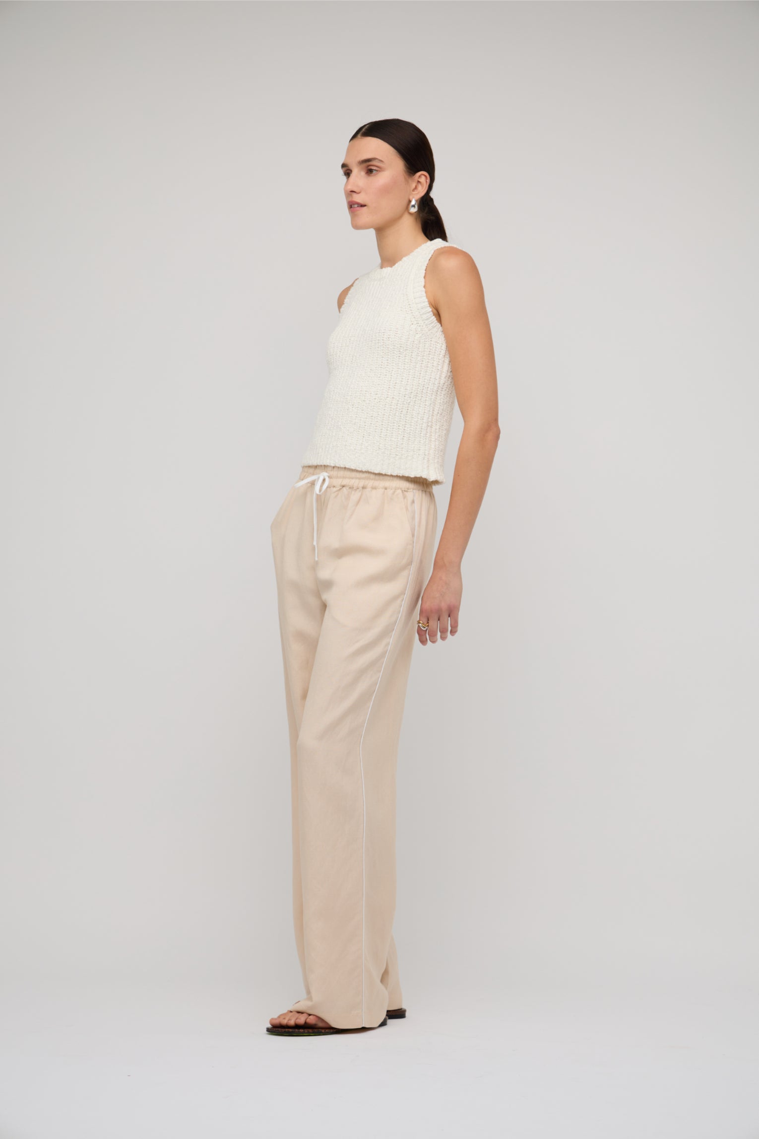 Woman wearing a white sleeveless top and beige pants on a plain background