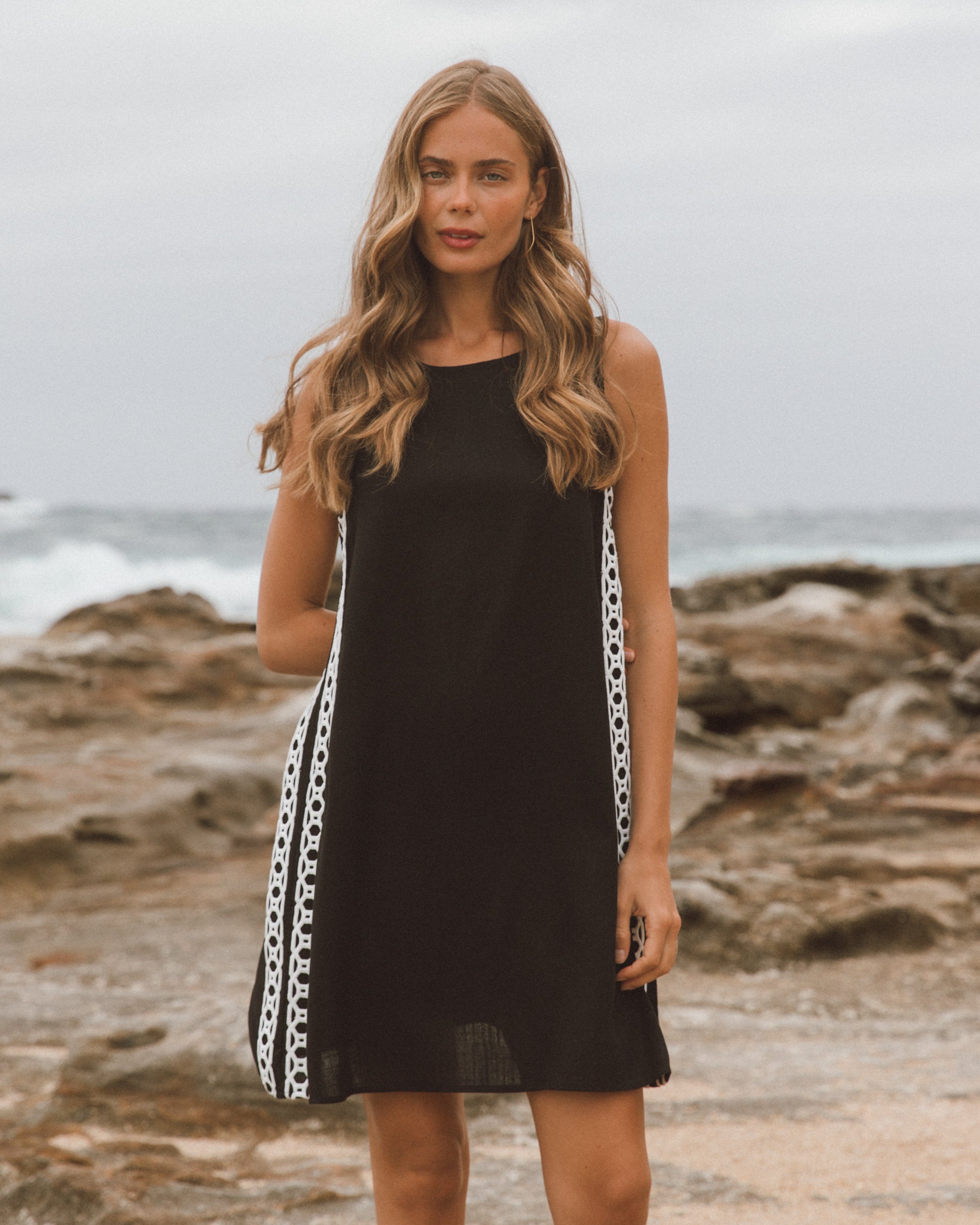 Woman in a black dress with chain detail standing on a rocky beach.