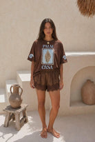 Woman wearing a brown 'Palm Casa' t-shirt and shorts in a sunlit outdoor setting.