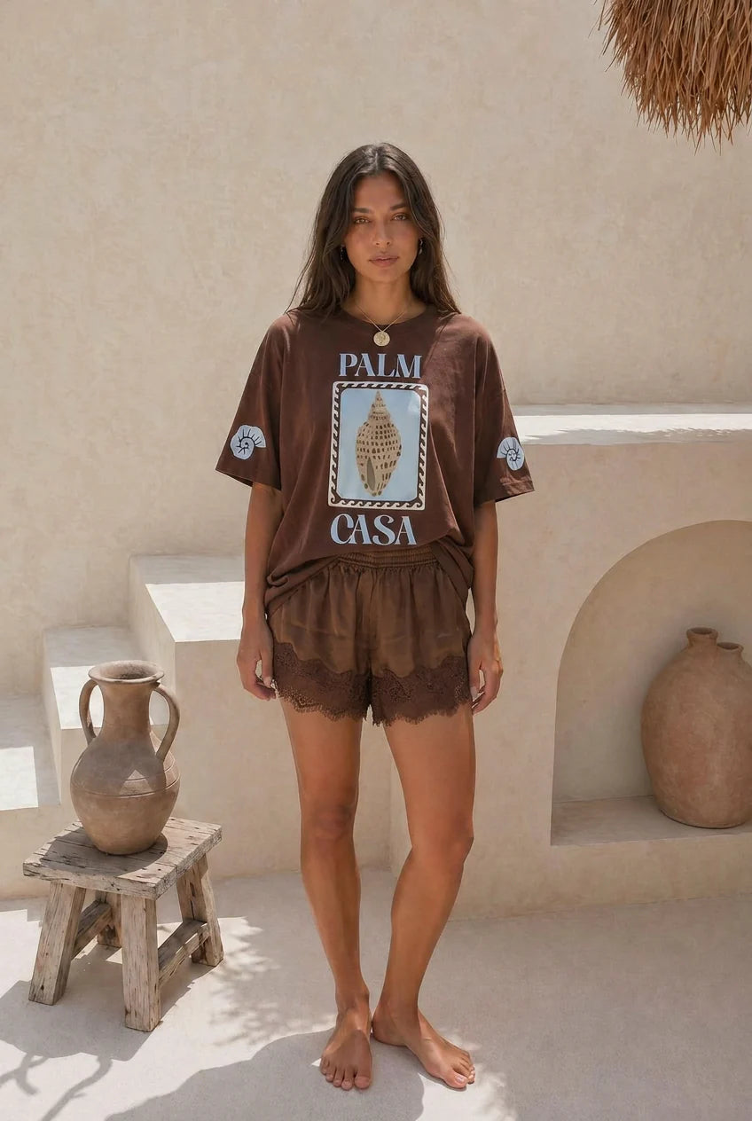 Woman wearing a brown 'Palm Casa' t-shirt and shorts in a sunlit outdoor setting.