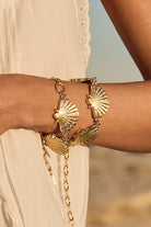 Gold bracelet with shell designs on a person's wrist against a blurred natural background