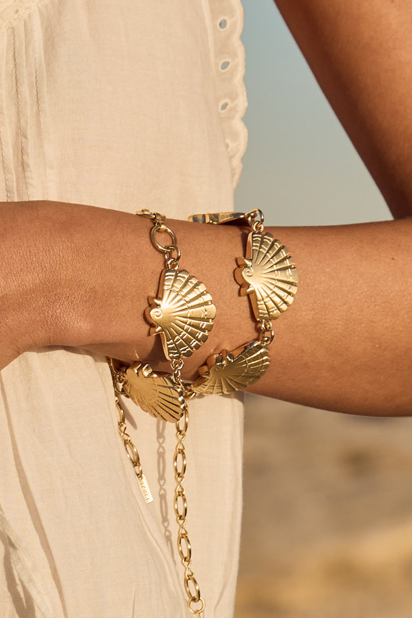 Gold bracelet with shell designs on a person's wrist against a blurred natural background
