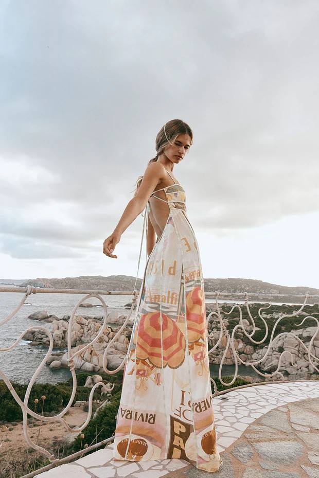 Woman in a floral dress standing by a scenic coastal area with a cloudy sky.