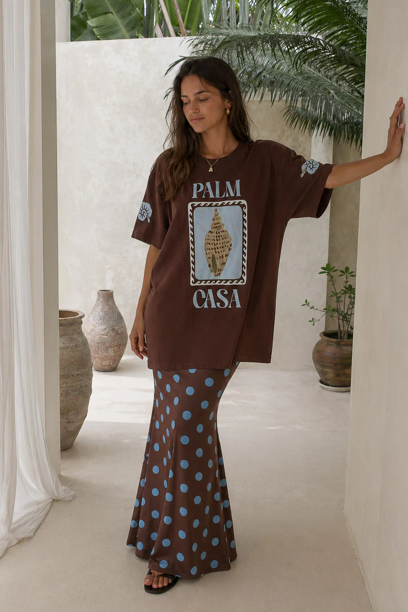Woman wearing a brown oversized t-shirt with 'Palm Casa' design and patterned skirt in an outdoor setting.