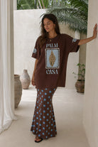 Woman wearing a brown oversized t-shirt with 'Palm Casa' design and patterned skirt in an outdoor setting.