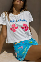 Person wearing a white t-shirt with 'Copacabana' and floral designs, sitting against a beige wall.