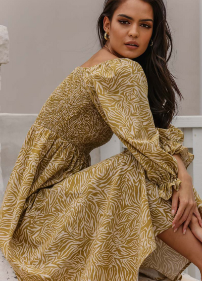 Woman sitting by a wall wearing the animal print khaki and neutral paros dress with gold accessories
