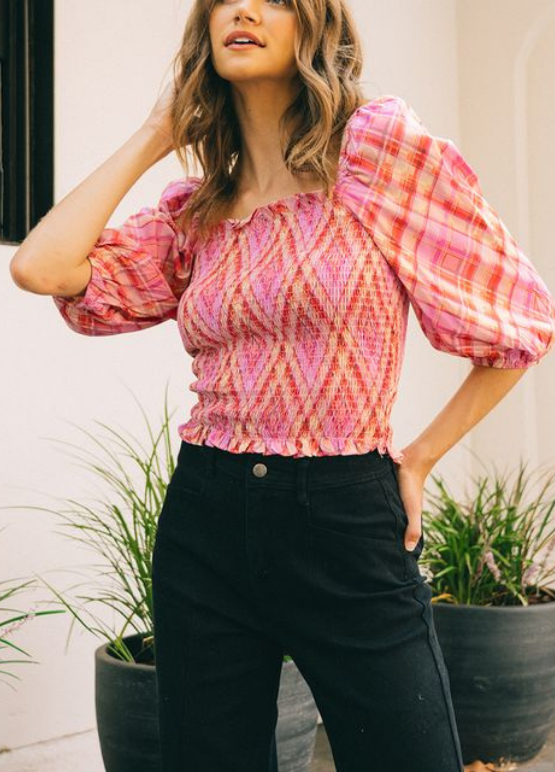 A woman wearing a pink and red checkered top with mid-length sleeves, styled off the shoulder, paired with black pants.